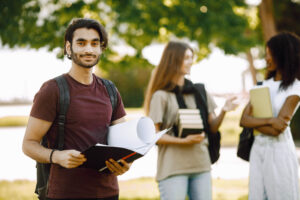 Joven estudiante de origen colombiano o mexicano con libros en mano, de pie en un parque universitario en Canadá, preparándose para planear su residencia permanente.