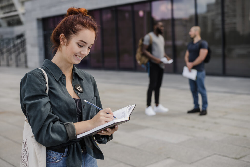 Mujer sonriente escribiendo en un cuaderno mientras planifica su futuro de residencia permanente en Canadá, con dos compañeros conversando en el fondo.