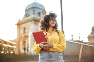 Estudiante universitaria con gafas y carpeta roja en la mano, sonriente, en un campus histórico.